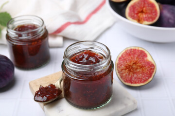 Glass jars of tasty sweet fig jam and fruits on white tiled table