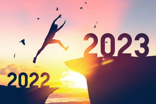 Silhouette man jumping between cliff with number 2022 to 2023 and birds flying at tropical sunset beach. Freedom challenge and travel adventure holiday concept.