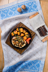 Pork with fried mushrooms, potatoes, zucchini, tomatoes and onions in a cast iron pan on a table.