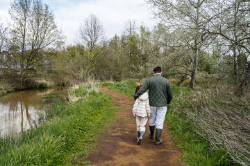 Naklejka na ściany i meble A father and a 10 years daughter walking on a path in the fields and swamps. They are hugging each other, and have outdoor outfit and rain boots on.