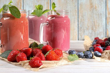 Mason jars of different berry smoothies and fresh ingredients on white wooden table