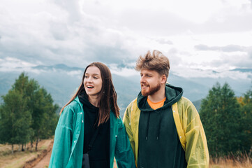 Photo of a happy couple of tourists on a hike standing on the top of a mountain wearing raincoats and looking away with a smile on their face