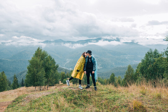 Cute couple of tourists in raincoats standing in the mountains, hugging and kissing during the sunset on the background of cloudy mountain views