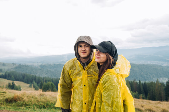 Beautiful young couple man and woman in raincoats standing with smiles on their faces against the background of mountain views and posing for the camera with a serious face. - Powered by Adobe