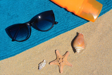 Fototapeta na wymiar Stylish sunglasses, bottle of sunblock and blue towel on sand, flat lay. Beach accessories