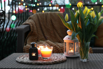 Soft blanket, burning candles and yellow tulips on rattan garden furniture in evening