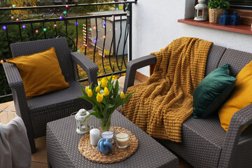 Soft pillows, blanket, candles and yellow tulips on rattan garden furniture outdoors