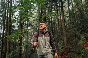A handsome male hiker with a backpack and a stick in his hand is walking in the mountains through a dense forest with a serious face and looking away. Active tourism.м