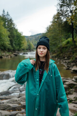 A beautiful tourist girl in casual clothes and a raincoat stands on the background of a mountain river and beautiful views of the forest and looks at the camera with a serious face.