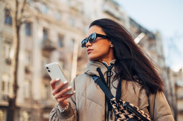 Beautiful brunette woman in sunshine stands on the street with a smartphone in hand and looks away with a serious face.
