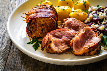 Medium well done pork loin wrapped in bacon with fried potatoes and feta cheese on beets on wooden table
