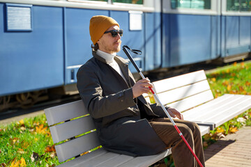 Blind man with a walking stick sits on a bench on the background of a city tram