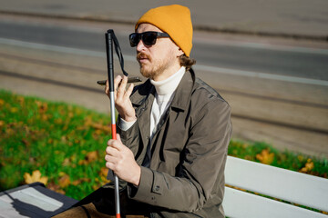 Blind man with a walking stick sits on a bench uses a smartphone