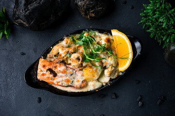 Baked red fish salmon with mushrooms, potatoes, cheese and sauce, garnished with herbs and lemon on a dark backgound.