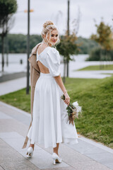 Fototapeta na wymiar A beautiful blonde bride with a bouquet of wild flowers with reeds walks in autumn with a beige coat, looking back. Wedding photography, portrait.