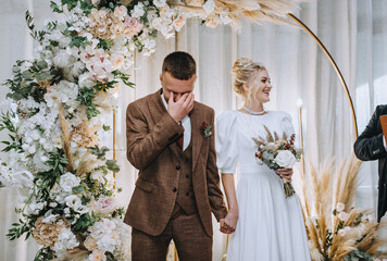 Stylish groom in brown suit wipes tears with hand, cries while standing with beautiful smiling...