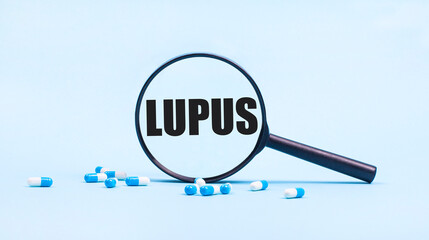 On a blue background, white and blue capsules with pills and a black magnifying glass with the text LUPUS. Medical concept.