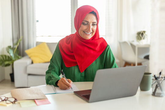 Happy mature muslim businesswoman taking notes while sitting with laptop and working online at home, free space - Powered by Adobe