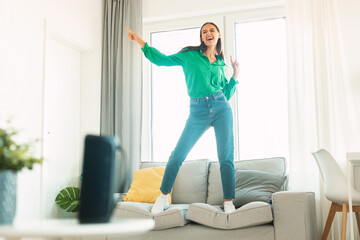 Happy woman listening to song via modern music speaker, having fun and dancing on sofa at home,...