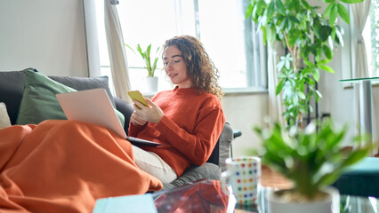 Relaxed young woman sitting on sofa holding mobile phone using cellphone and laptop technology doing online ecommerce shopping, texting messages relaxing on couch in cozy living room home interior.
