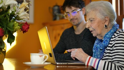 Closeup of elderly senior woman learning to use a laptop computer from teen male grandson with tea.