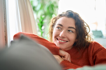 Young smiling pretty curly woman relaxing sitting on couch at home and dreaming. Happy relaxed calm beautiful lady enjoying comfort thinking or good on soft sofa looking away. Close up. - obrazy, fototapety, plakaty