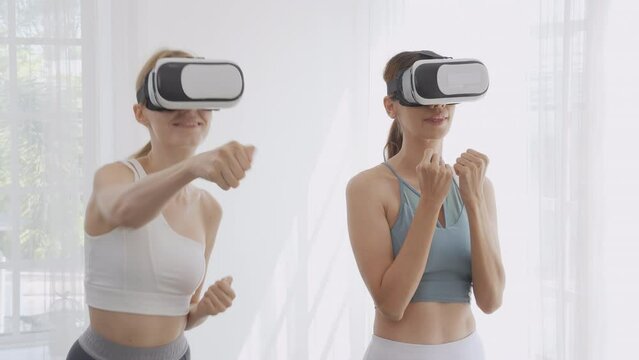 Two young caucasian woman wearing vr headset while workout and exercise together at living room, futuristic and innovation of virtual reality metaverse, female playing game and punch with enjoy.