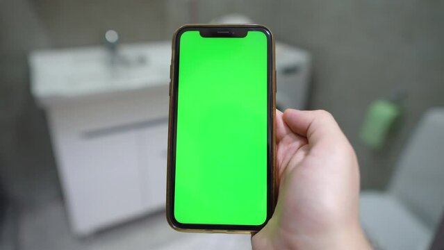 Use green screen for copy space closeup. indoors of toilet or bathroom. Chroma key mock-up on smartphone in hand.  young man holds mobile phone iPhone and swipes photos or pictures