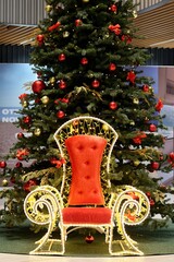 Fototapeta na wymiar Throne for Santa Claus under the Christmas tree. Colorful balls garland glowing lamps and red berries on the branches are sprinkled with snow.Festive traditional seamless background for the New Year