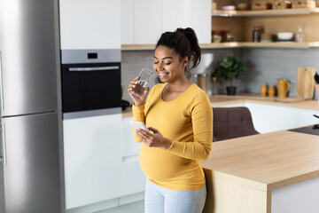 Cheerful young black pregnant lady with big belly, chatting on smartphone, drink glass of water