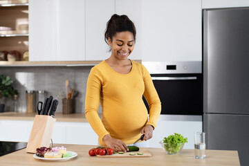 Glad young black pregnant woman with big belly cuts organic vegetables, prepare salad