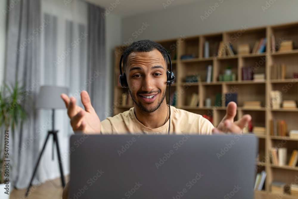 Poster happy arab man in headphones having video call on laptop, talking and gesturing at computer webcamer