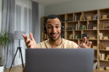 Happy arab man in headphones having video call on laptop, talking and gesturing at computer...