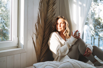 Young woman with blonde curly hair in white cardigan with cup of cocoa in hands looking at window...