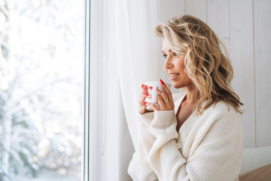 Young woman with blonde curly hair in white cardigan with cup of cocoa in hands looking at window with winter landscape at home
