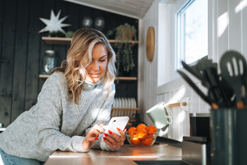 Young woman with blonde curly hair in grey sweater using mobile phone in hands in kitchen at house