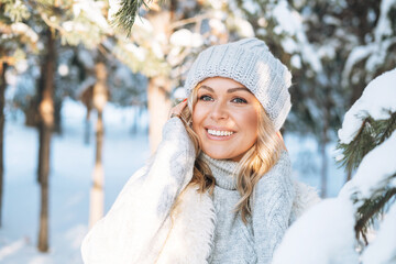 Close up portrait of smiling blonde woman with blonde hair in winter clothes hands near face against background of snowy winter forest