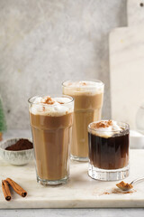 Three tall glasses with warm coffee drink with cinnamon, whipped milk foam and caramel on a marble board and christmas ornaments and decoration