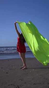 View From Behind Of Young Latin Woman Standing On The Shore Of The Beach Holding A Veil Or Cloth With Her Arms Above Her Head