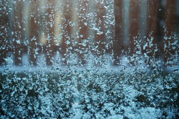 Glasscheibe mit Wasser- und Eismuster vor Gartenmauer und braunem Lattenholzzaun am Morgen im Winter