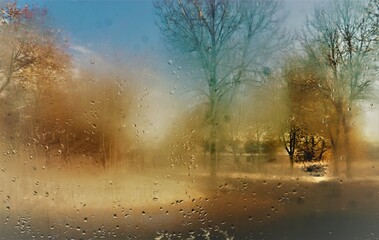 Feuchte beschlagene Glasscheibe mit Regentropfenmuster vor Schneelandschaft mit Bäumen und Himmel bei Sonne am Mittag im Winter