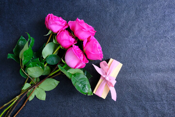 Pink roses, bouquet placed on a dark stone background. Next to it is a gift box wrapped in pink paper with a ribbon tied around it. The scene depicts holiday greetings as well as love and romance.