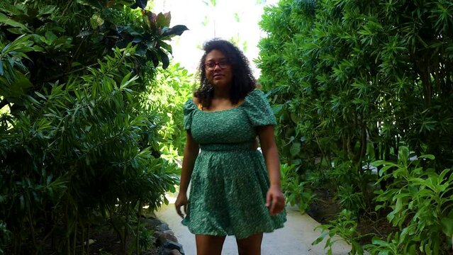beautiful black woman is walking down the narrow green garden pathway happily while wearing a pretty dress for the summer. 
