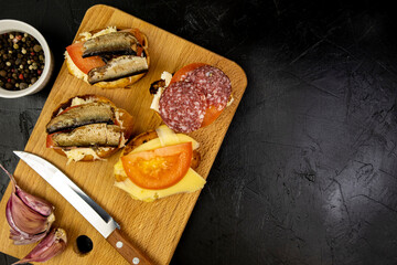 Light sandwiches with spices on a cutting board with fish sausage cheese and tomatoes on a black background top view, light snacks for beer