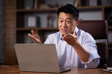 Fototapeta na wymiar Asian man holding online webinar using laptop webcam, sitting in a home office. Man having online meeting and communicating remotely, looking at computer screen