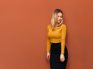 Portrait of beautiful young blonde with big breasts stands half-sideways, Lowering her hands to bottom, looking at her shoulder, in black dress and yellow blouse, isolated orange background.