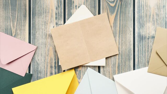 Looped stop motion video of white opening envelope on gray wooden background. Multicolored envelopes fall from above, kraft paper card is taken out of envelope and opened. Copy space. Top view