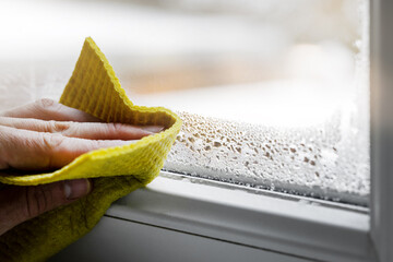 high humidity in the house. hand wipes off water condensation from plastic window glass in the room. home moisture - 547749466