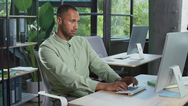 Portrait of successful confident businessman in casual outfit sitting at computer and typing on keyboard. Handsome African-American man showing thumbs up to camera in office. Indoors. Agree and