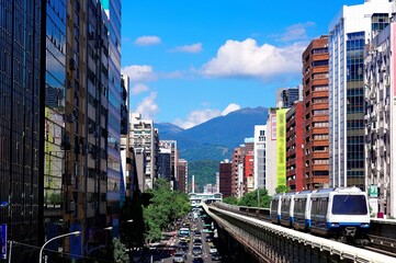 A metro train traveling on the elevated rail of Taipei MRT between office towers under blue sky ~...